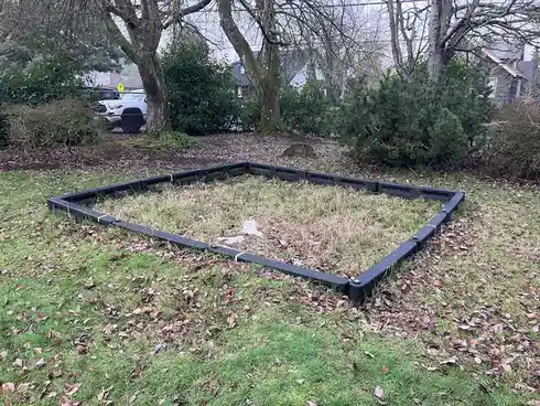 An outdoor photo shows a grassy yard scattered with fallen leaves, featuring a black plastic barrier fence forming an…