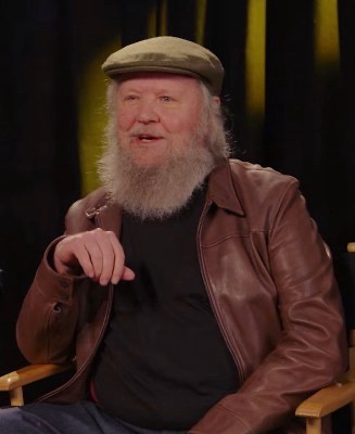 A man with a long white beard and a brown leather jacket sits in a chair, pointing his finger forward with a serious…