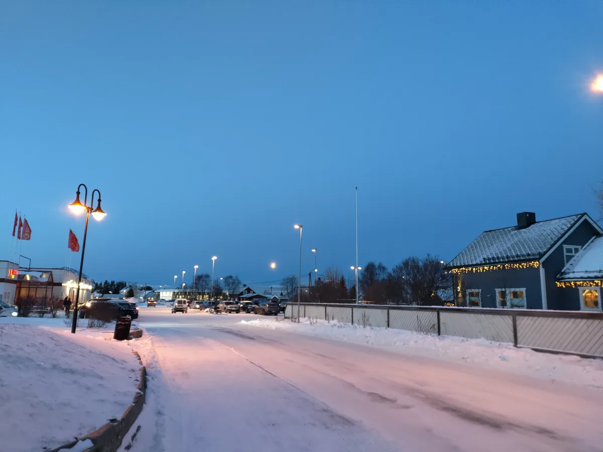 A snowy street at dusk with streetlights glowing, cars parked along the sides, and a house decorated with string lights. The…