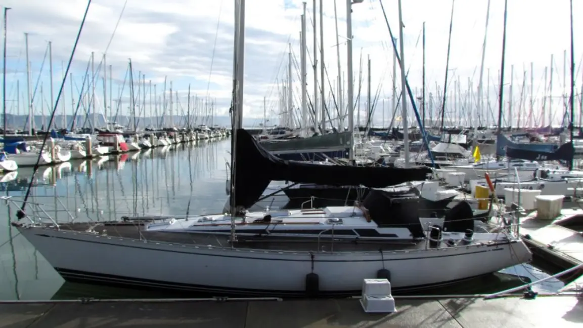 A white sailboat with its sails covered in black cloth is docked in a marina filled with other boats. The overlaid text…