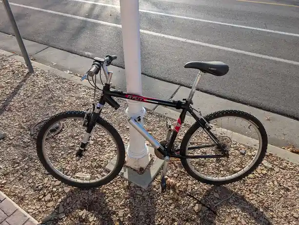 A black mountain bike with red accents is parked upright against a white pole on a gravel sidewalk next to a paved road. The…