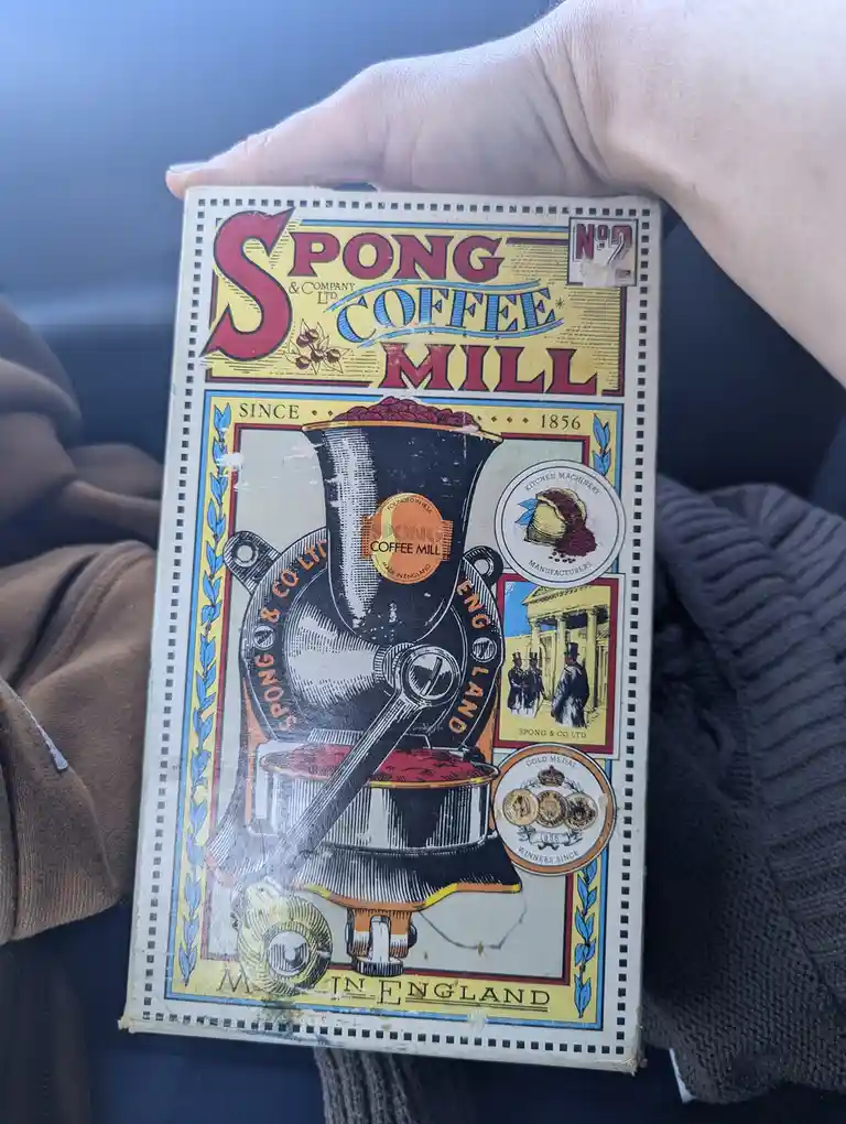 A hand holds a vintage-style box for a 'Spong & Company Ltd. Coffee Mill', featuring an illustration of an old-fashioned…