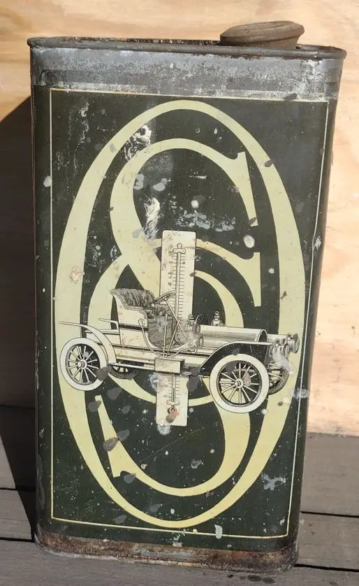 A weathered, vintage metal fuel can with a faded green and cream label depicting an early 20th-century automobile inside a…