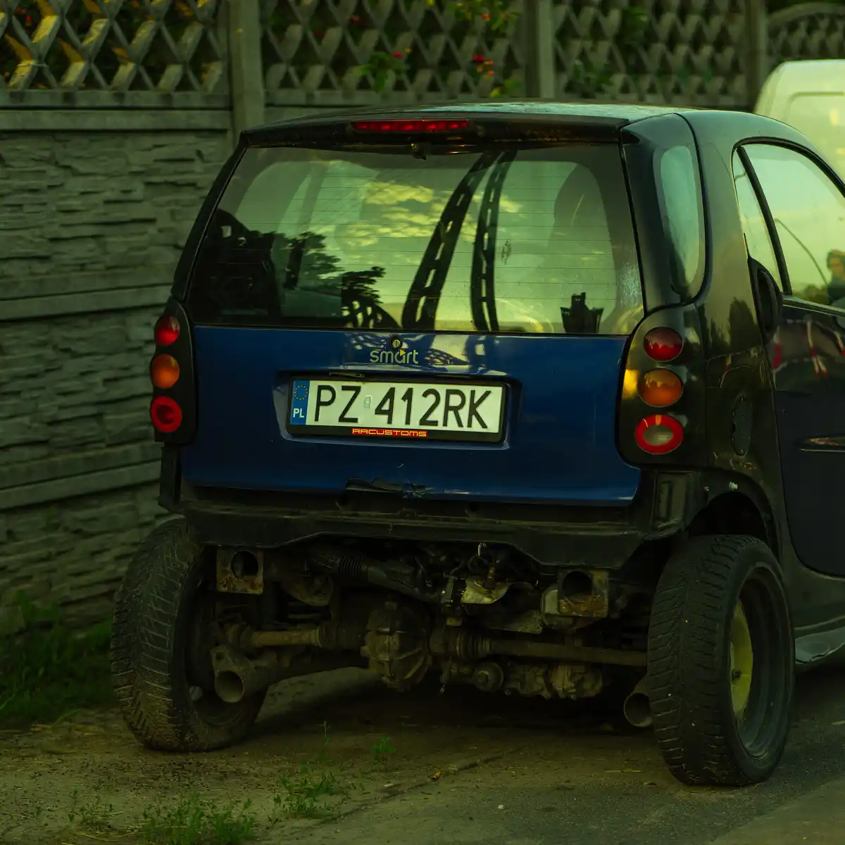 The image shows the rear of a blue Smart car parked outdoors, with its entire undercarriage exposed and visibly rusted…