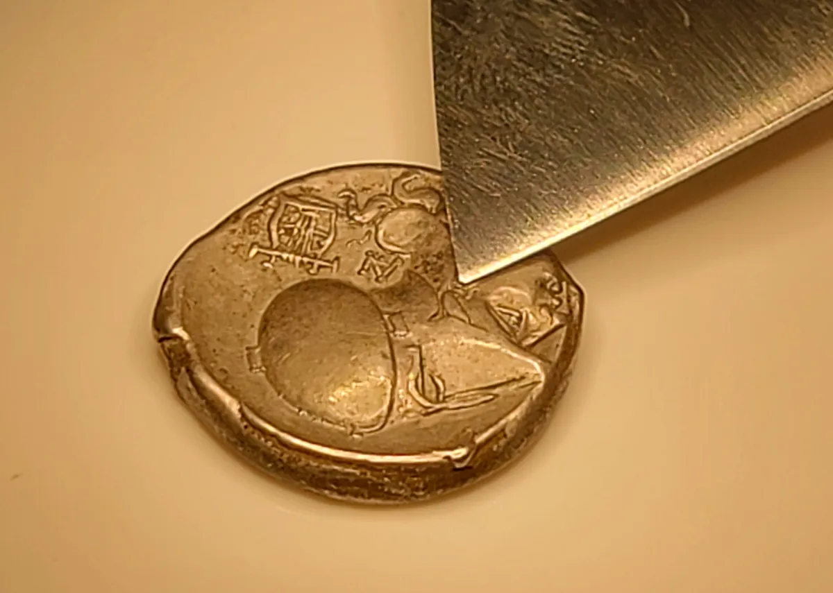 A close-up photo shows a worn, irregularly shaped metallic coin with embossed designs, partially pierced by the blade of a…