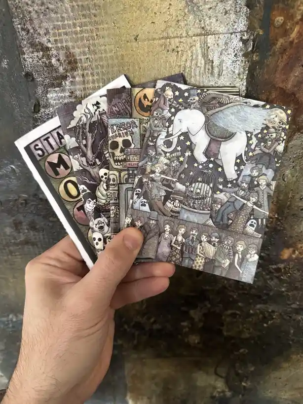 A hand holds a fan of five illustrated cards against a rusty metal background. The cards feature dark, whimsical, and gothic…