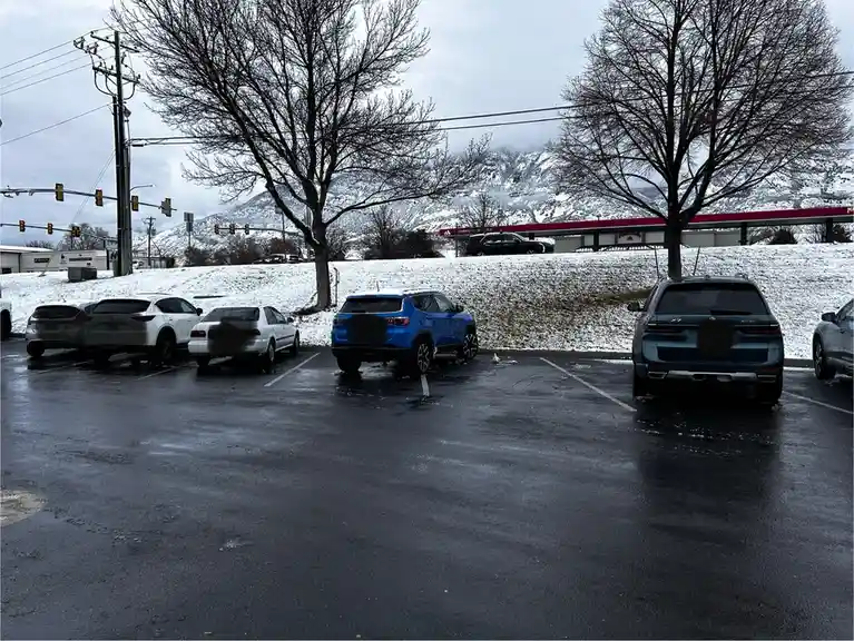 A wet parking lot with several cars parked, including a blue SUV and a teal SUV, with snow on the ground and snow-covered…