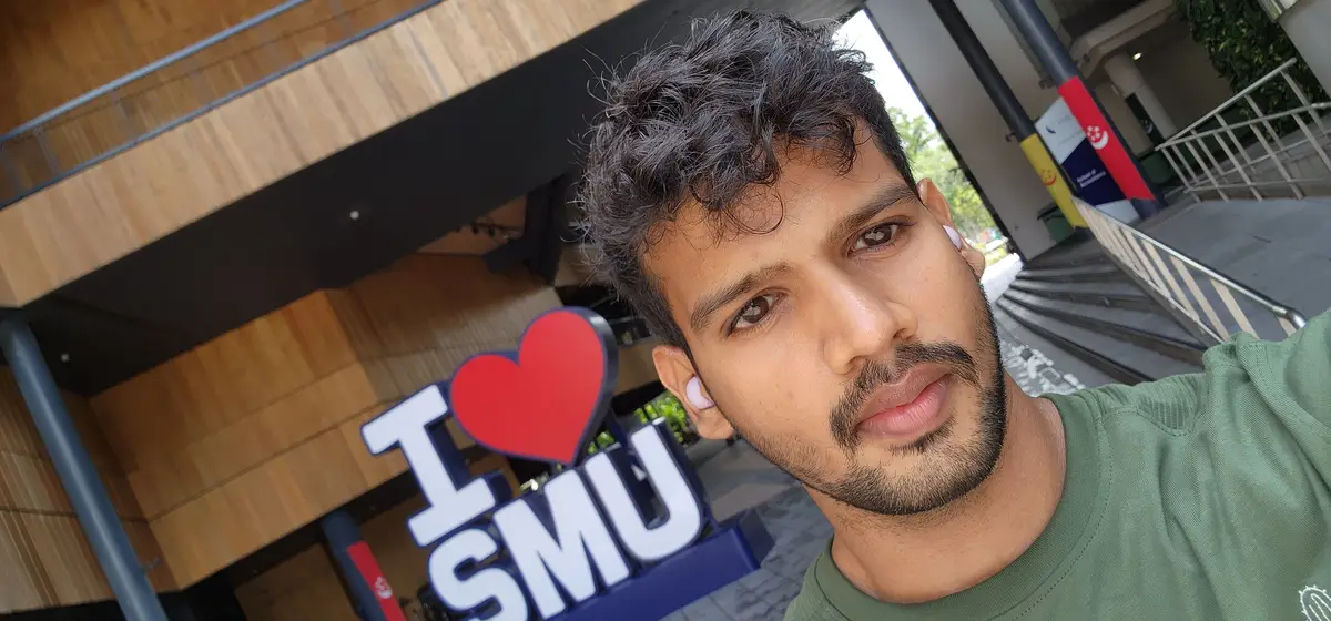 A young man with dark curly hair and a green shirt takes a selfie in front of a large 'I ❤️ SMU' sign, which features a red…
