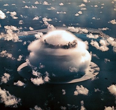An aerial view shows a large, white, mushroom-shaped cloud formation hovering over a ring of land surrounded by ocean, with…