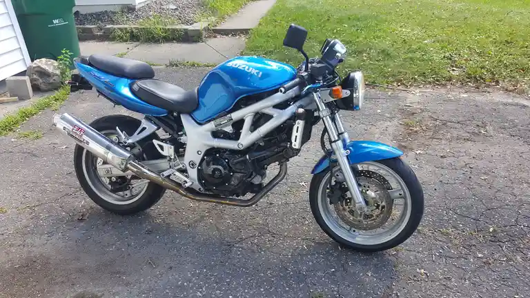 A blue Suzuki SV650 motorcycle is parked on an asphalt driveway next to a grassy lawn and a green waste management bin. The…