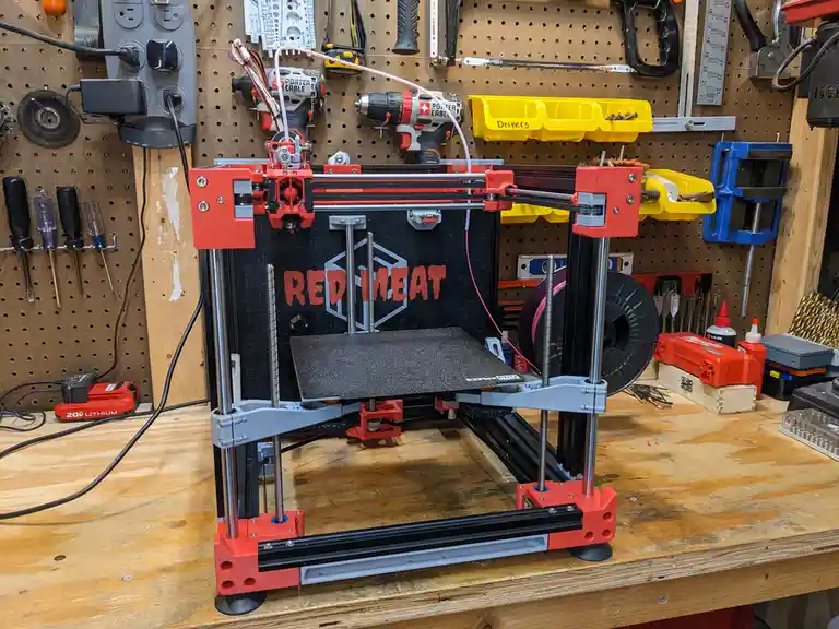 A red and black 3D printer sits on a wooden workbench in a cluttered workshop, with tools like drills, screwdrivers, and…