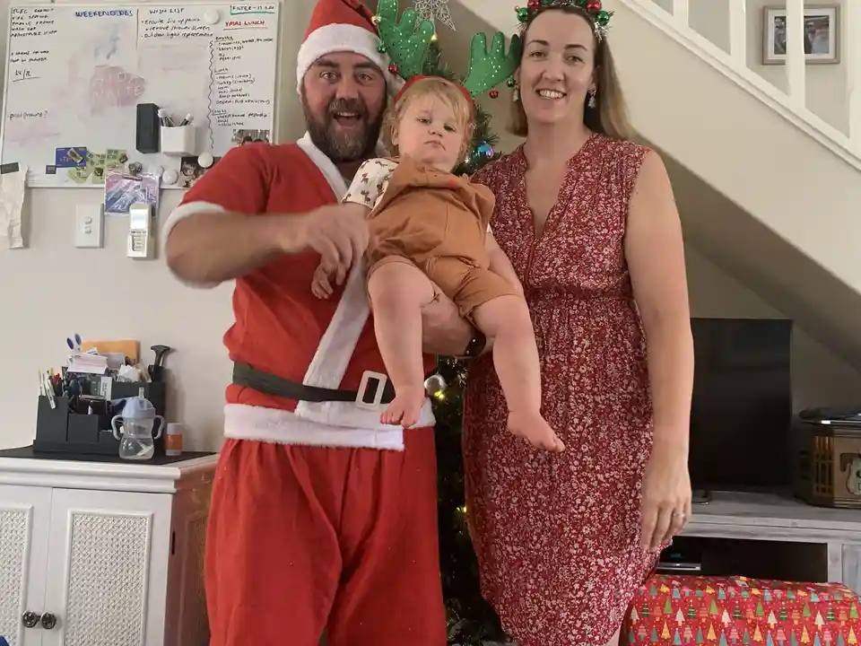 A man dressed as Santa Claus, wearing a red suit and green reindeer antlers, holds a baby who looks unimpressed. Beside…