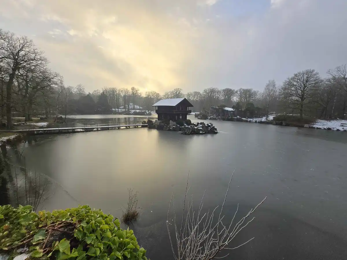 A serene winter scene shows a frozen lake with a small wooden house on a rocky island, snow dusting the trees and roof, and…