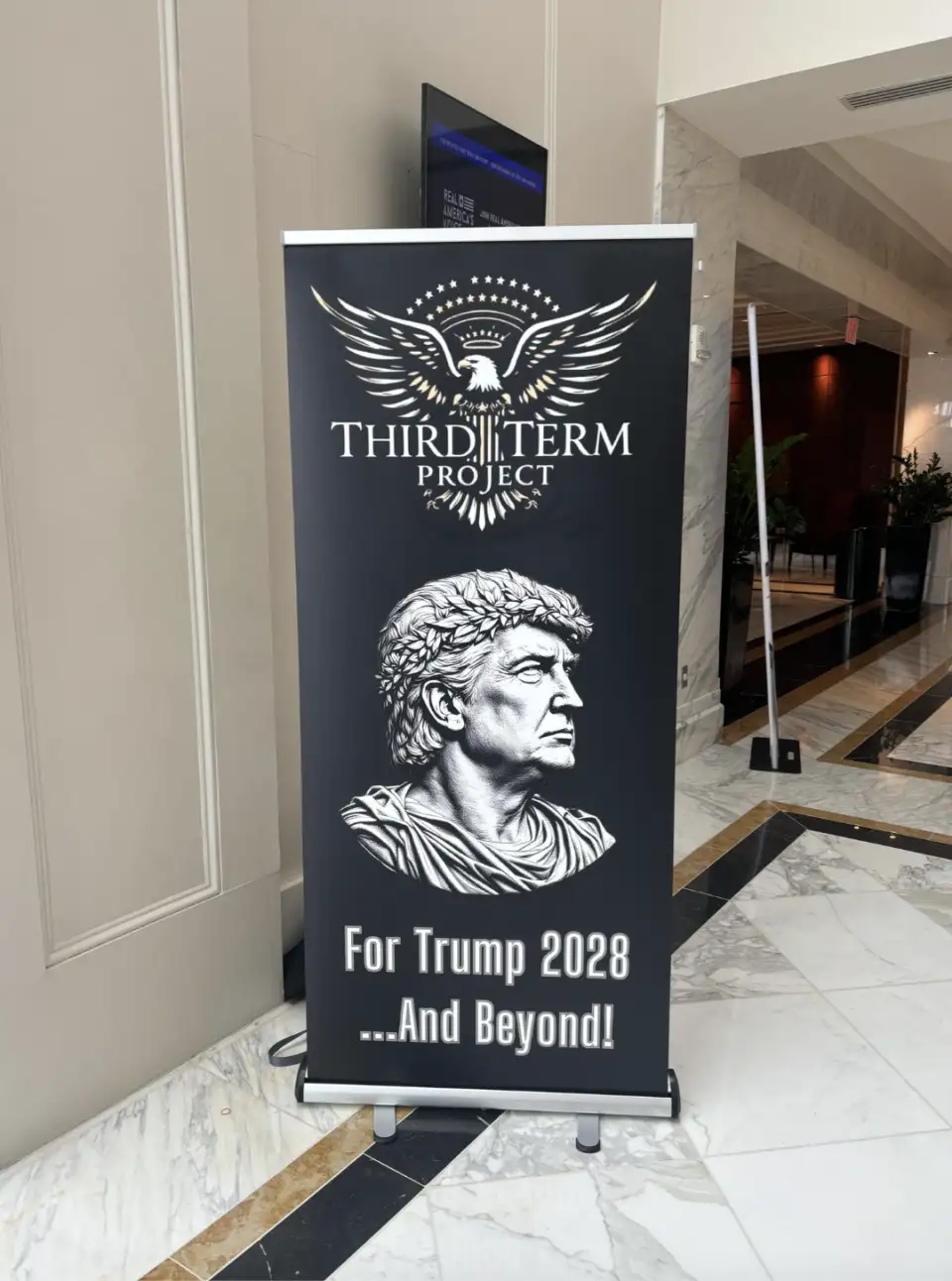 A black banner stand in a marble-floored lobby displays a logo of an eagle with stars above the text 'THIRD TERM PROJECT'…