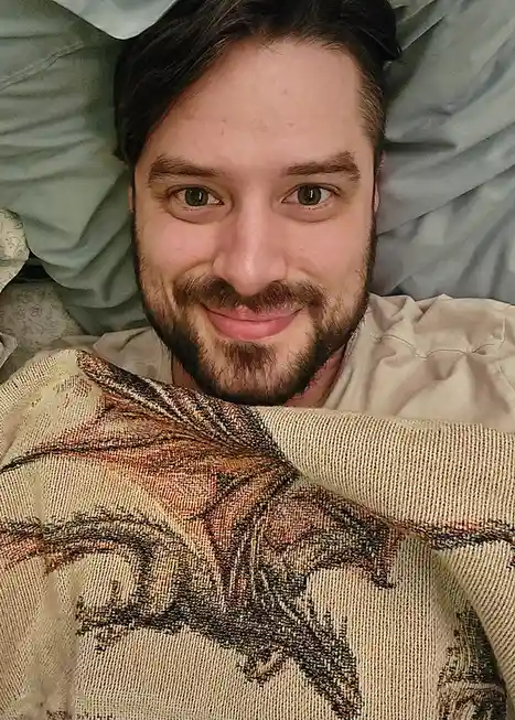 A man with dark hair and a beard lies in bed under a patterned blanket, smiling at the camera. He is wearing a light-colored…