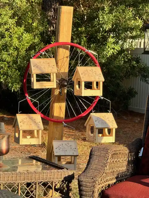 A red bicycle wheel mounted vertically on a wooden post has five small wooden birdhouses hanging from its spokes and rim…