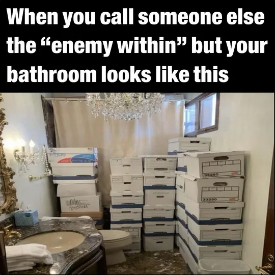 A bathroom is filled with stacks of white moving boxes, obscuring most of the space around a toilet and sink. The room has a…