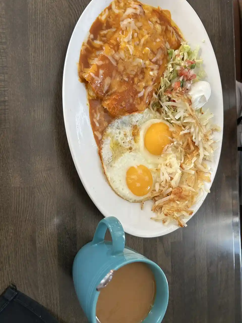 A top-down photo of a breakfast plate with two sunny-side-up eggs, enchiladas covered in sauce and cheese, hash browns, pico…