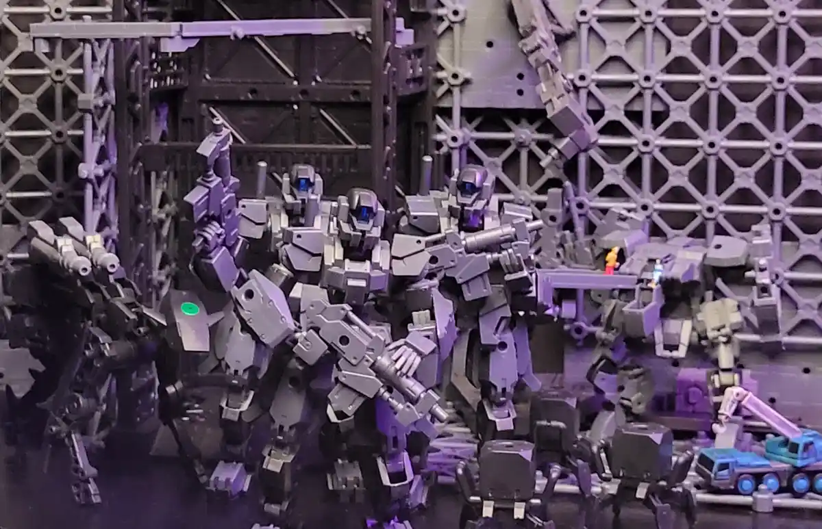 A diorama featuring multiple grey, heavily armed robot figures standing in a factory-like setting with metal scaffolding and…