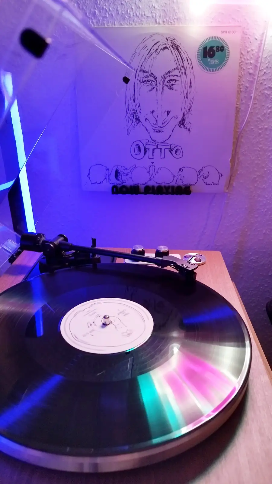 A vinyl record spins on a turntable under colorful lighting, with a custom album cover visible behind it. The cover features…