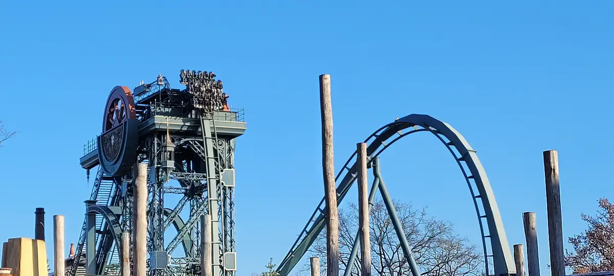 A roller coaster with a tall, ornate tower and a large loop against a clear blue sky. Riders are visible at the top of the…