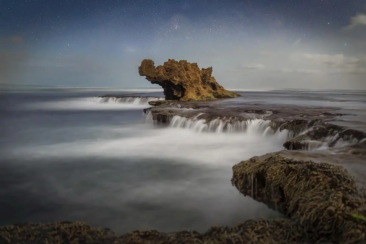 A long-exposure photograph captures a starry night sky above a rocky coastline where water flows smoothly over rocks…