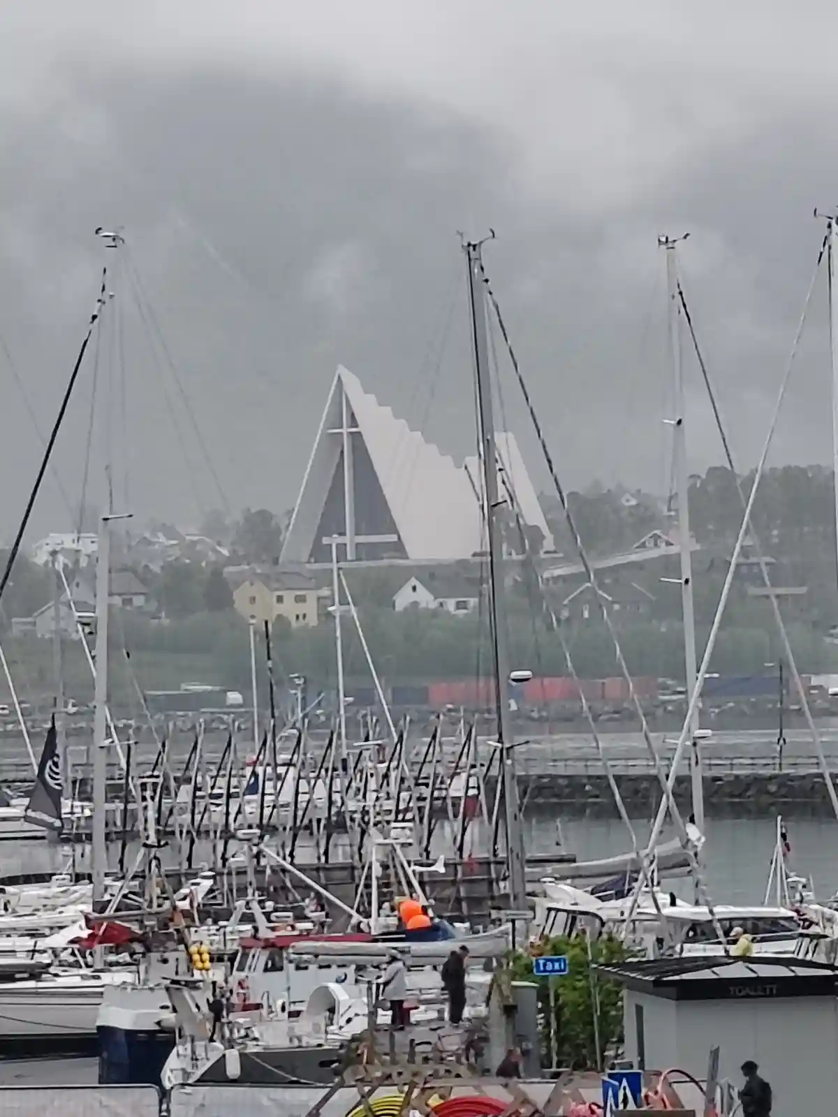 A photo of a harbor filled with sailboats and masts, with a distinctive white, angular church building visible in the…
