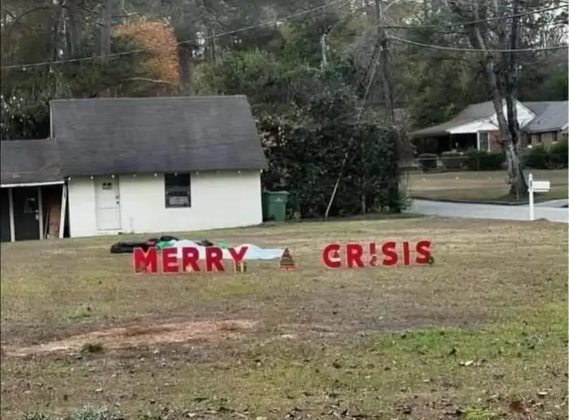A grassy yard in front of a small white house has large red letters spelling 'MERRY' and 'CRISIS' with a small Christmas…