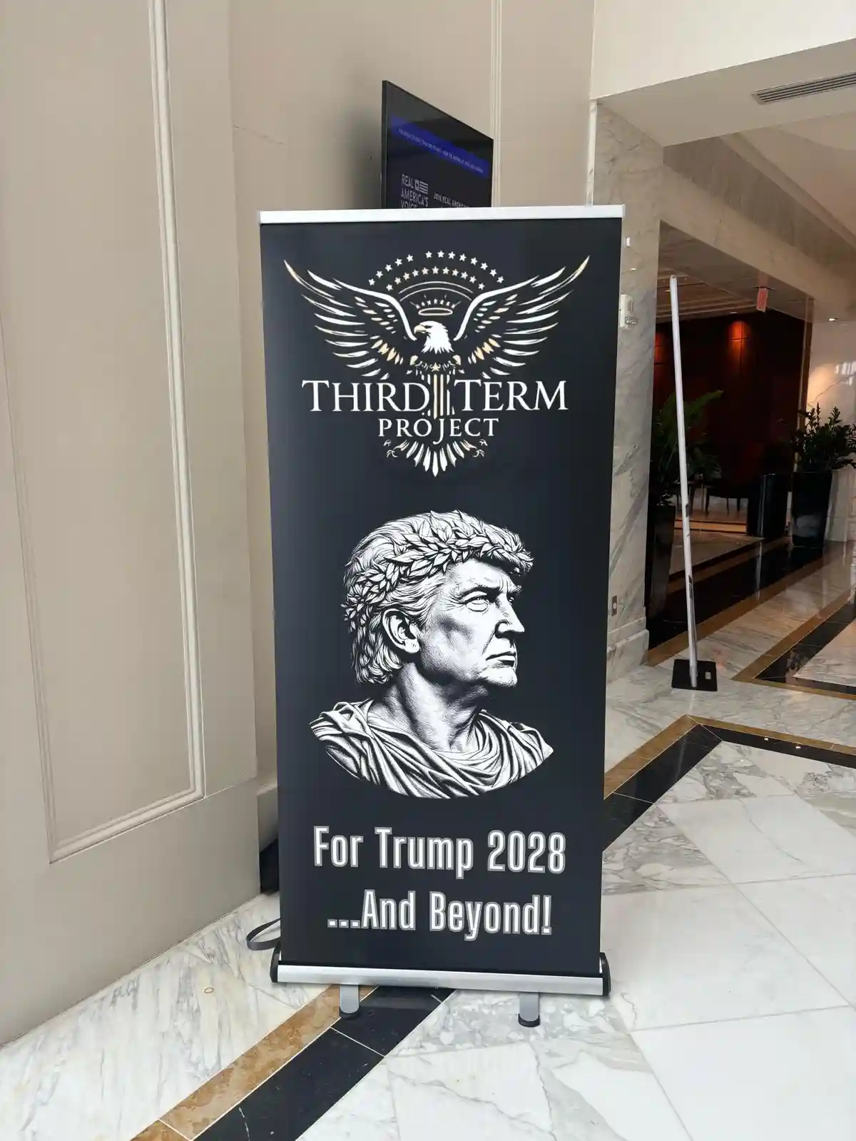 A black retractable banner stands in a marble-floored lobby, featuring a white graphic of an eagle with stars above the text…