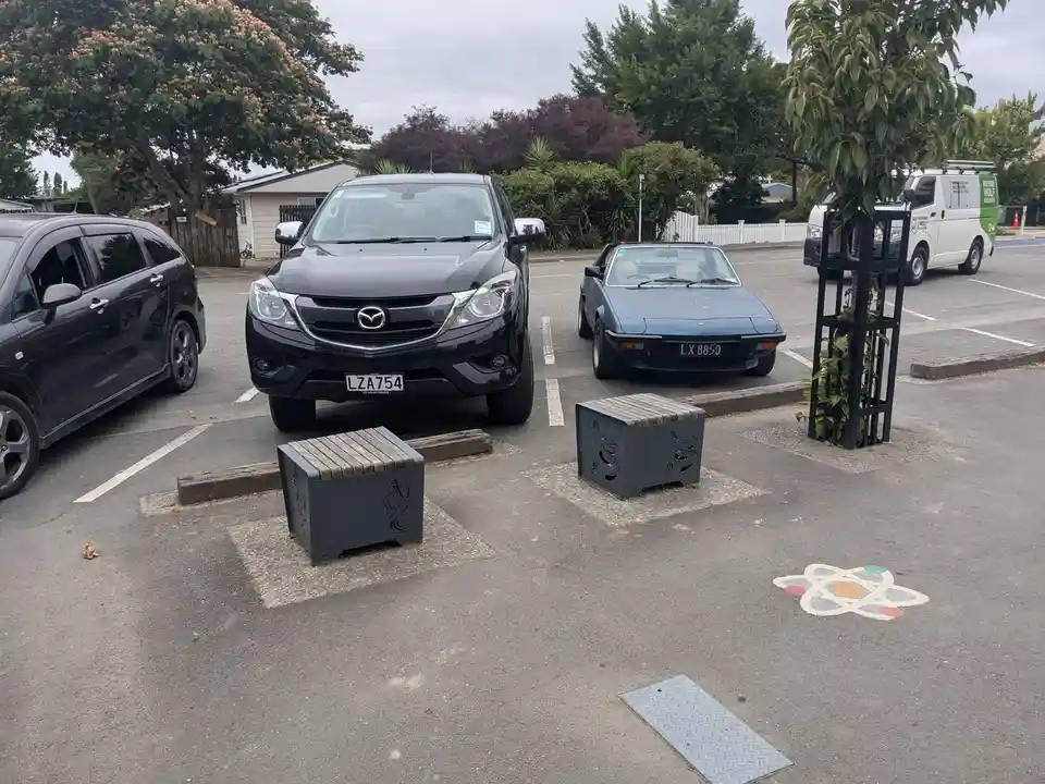 A black Mazda SUV and a blue classic convertible are parked in a lot with two modern, boxy public benches in front of them…