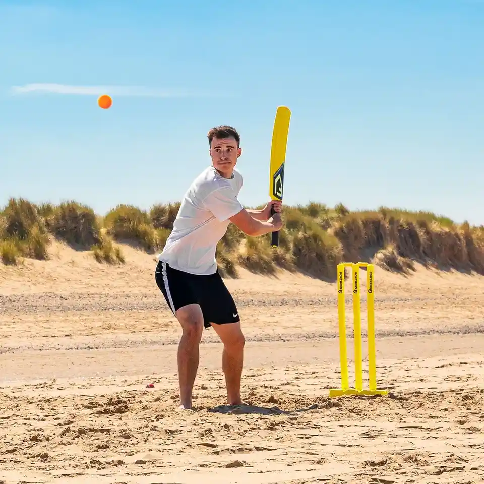 A young man stands on a sandy beach, holding a yellow cricket bat, poised to hit an orange ball that's in mid-air. In front…