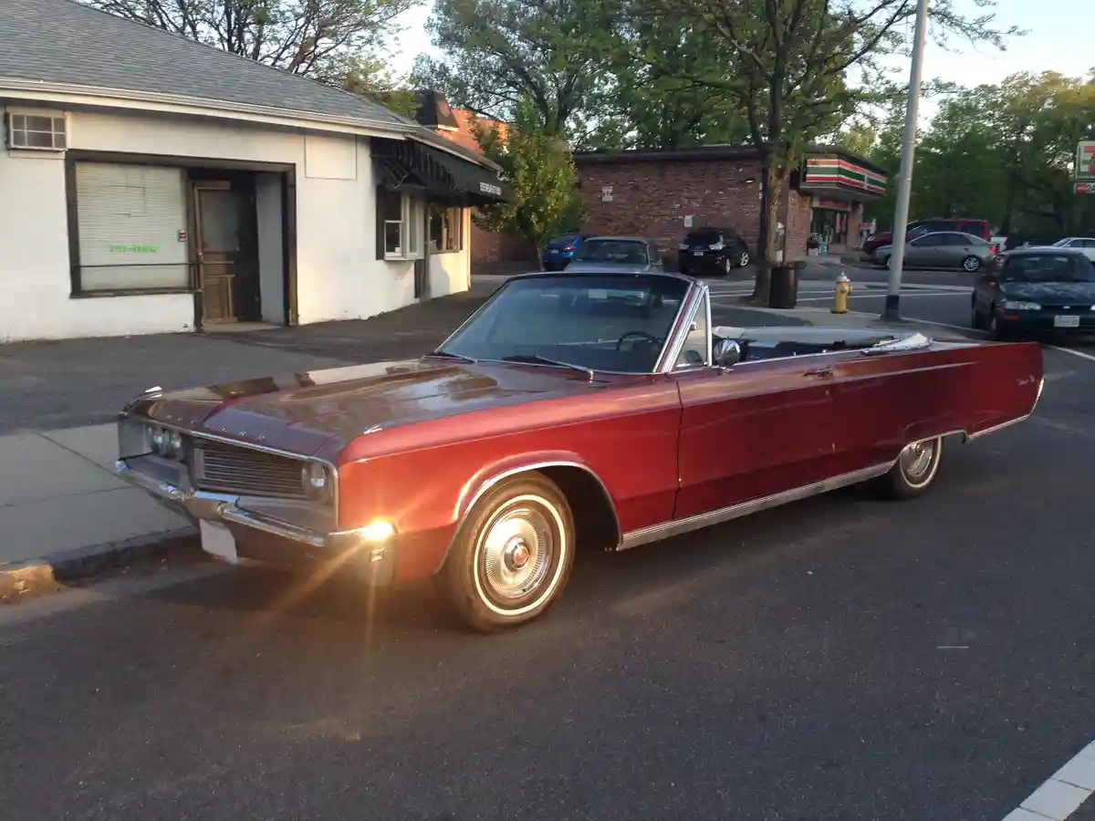 A shiny red 1960s Chrysler convertible is parked on a street in front of a 7-Eleven and other buildings. The car's top is…