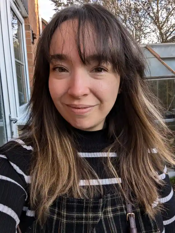 A woman with long brown hair and bangs smiles gently at the camera while wearing a black and white striped sweater and plaid…