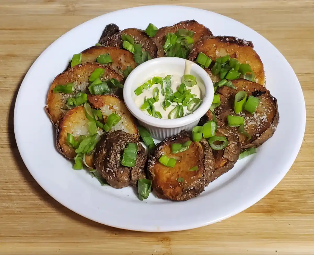 A white plate holds roasted potato slices arranged around a small bowl of creamy dip, all topped with chopped green onions…