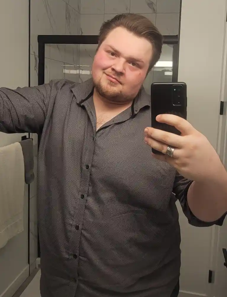 A man with a beard and short brown hair takes a mirror selfie in a bathroom wearing a dark grey button-up shirt, holding a…