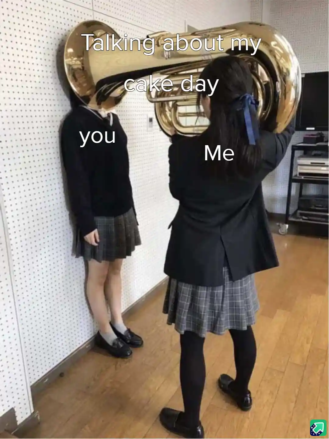 Two girls in school uniforms stand in a room; one holds a large brass tuba, obscuring her face, while the other stands…