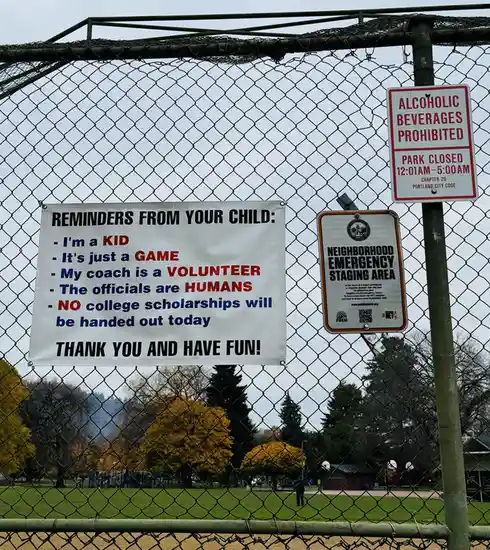 A white banner taped to a chain-link fence at a park reads 'REMINDERS FROM YOUR CHILD' with bullet points like 'I'm a KID'…