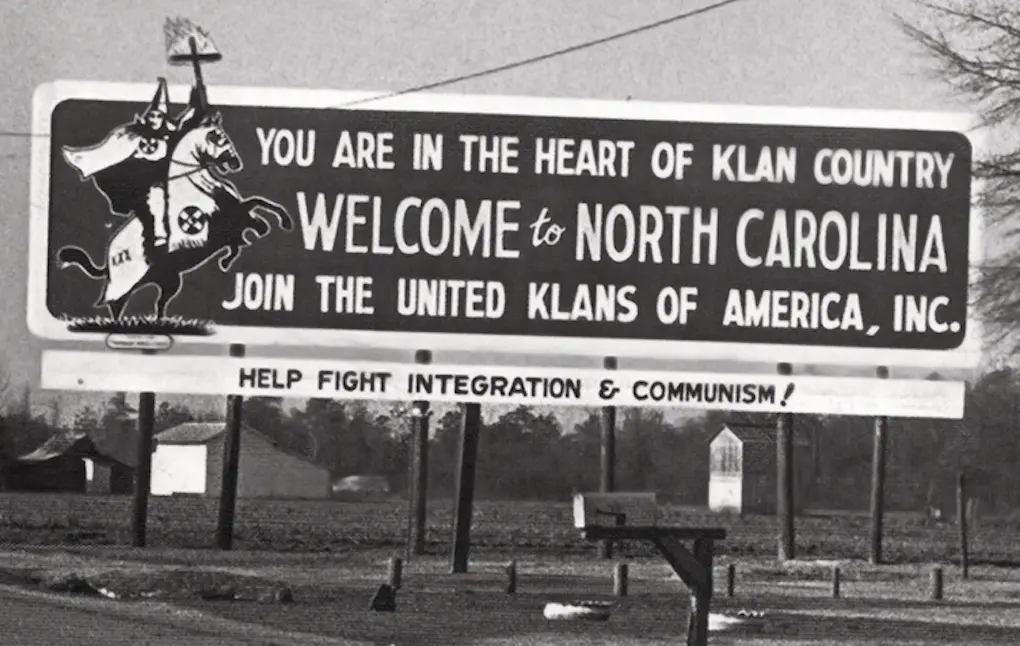 A black-and-white photo of a large roadside billboard from the mid-20th century. The sign features a graphic of a white-clad…
