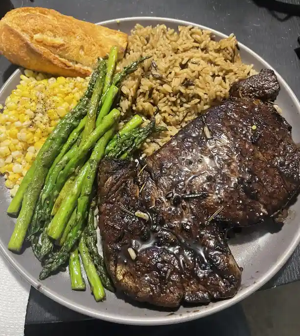 A close-up photo of a plated meal featuring a large, seared steak with sauce and herbs, alongside roasted asparagus…