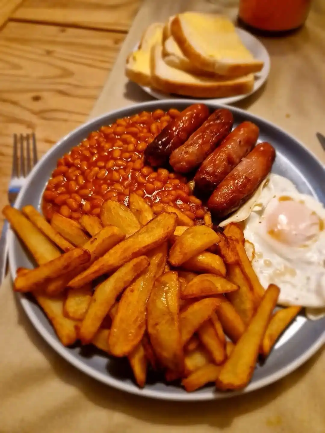 A close-up photo of a large plate of breakfast food including baked beans, four sausages, fried eggs, and thick-cut fries…