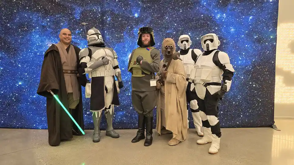 Six people dressed as Star Wars characters — a Jedi with a green lightsaber, a clone trooper, a stormtrooper, a Wookiee, and…