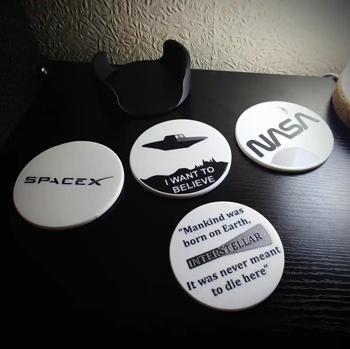 Four white circular coasters sit on a dark wooden table. One has the SpaceX logo, another the NASA logo, a third shows a UFO…