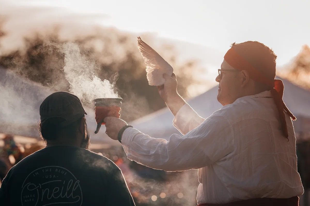 A man in a white shirt and red headband holds a smoking pot in one hand and a white bird in the other, seemingly performing…