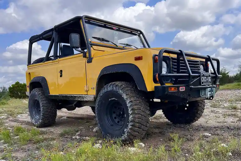 A yellow Land Rover Defender with large off-road tires and a black front bumper is parked on a dirt path with grass and…