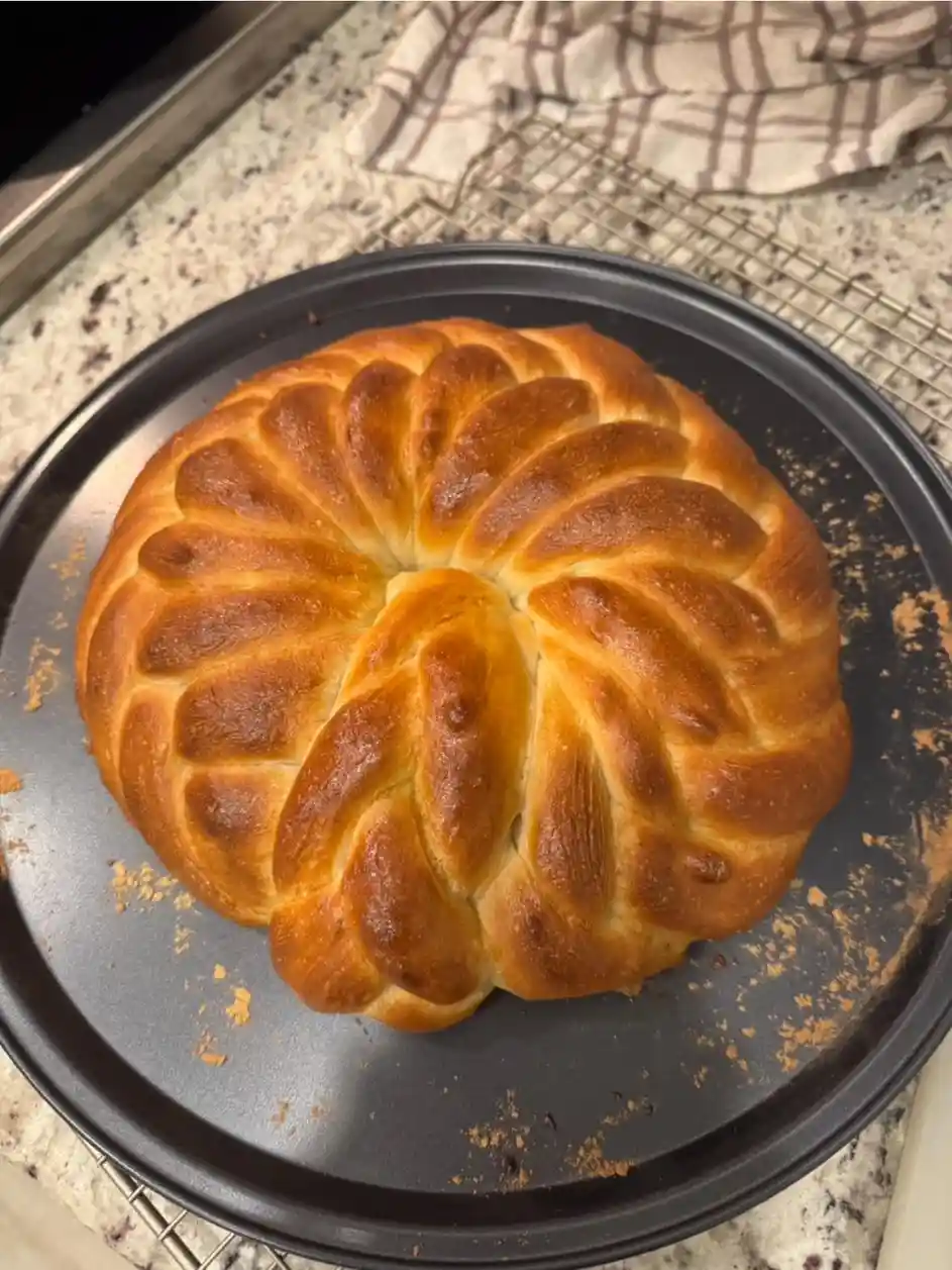 A golden-brown braided bread with a flower-like pattern sits on a black baking pan, resting on a wire cooling rack atop a…
