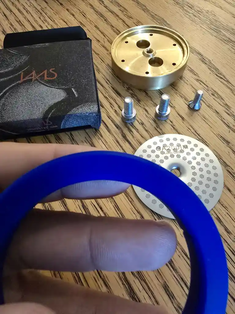 A hand holds a blue rubber band in the foreground, obscuring part of a wooden table. Behind it are several small metal parts…