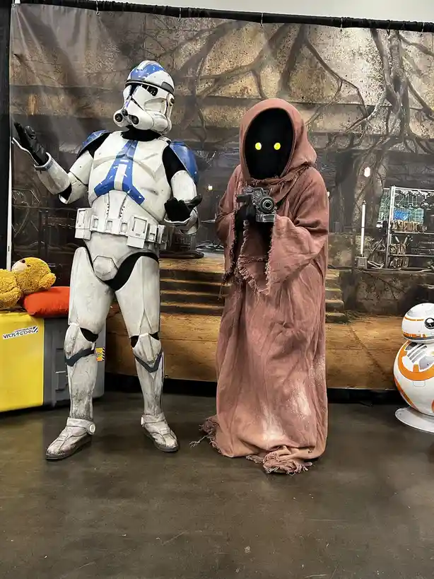 Two people in Star Wars costumes stand side by side: one as a Clone Trooper with white and blue armor gesturing with both…
