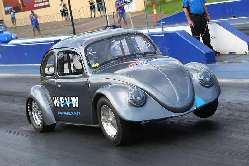 A silver, modified Volkswagen Beetle race car with 'WPW' and 'www.wpww.com.au' on its side is on a drag strip, angled as if…