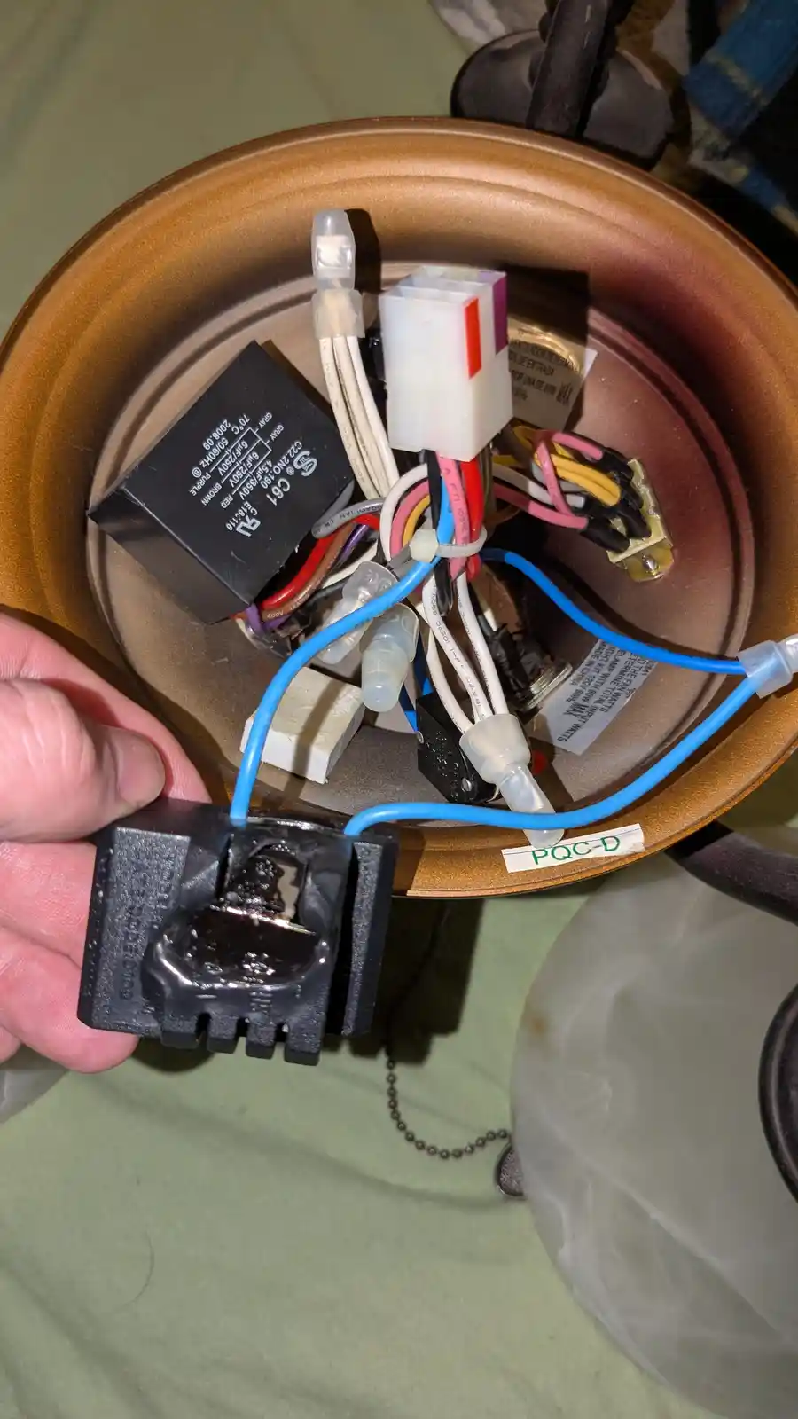 A hand holds a severely burnt and melted black electrical switch component, with visible charred residue and melted plastic…