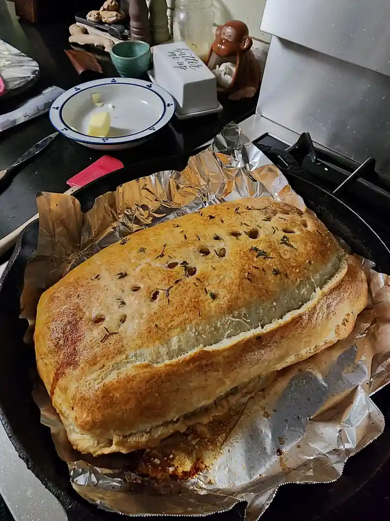 A golden-brown, rectangular loaf of bread with herbs and holes sits in a black cast-iron skillet lined with foil. In the…